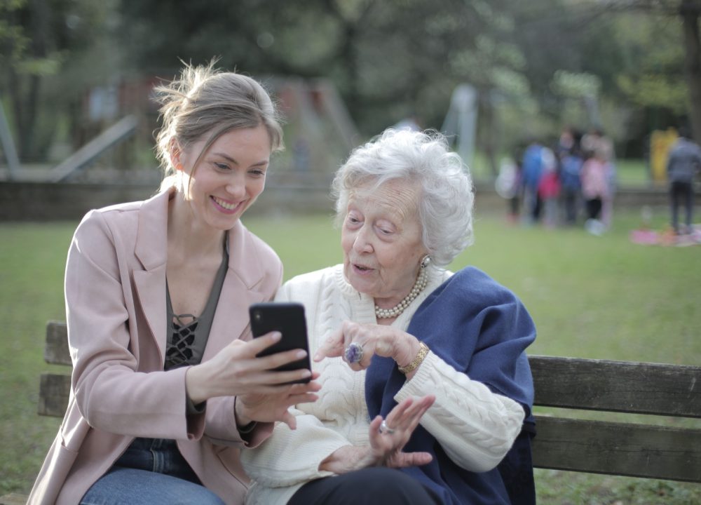 An adult daughter smiling with her senior mother, looking at a phone together on a park bench